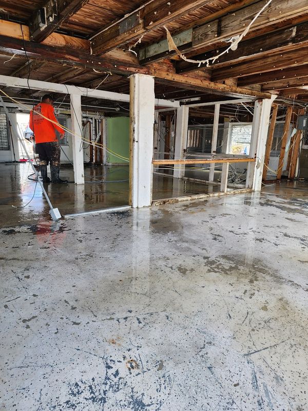 AHCM worker in safety gear pressure washing a flooded, storm-damaged building interior